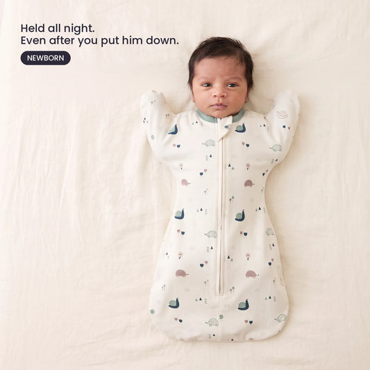 LIOR Held Swaddle Sack in Cream Meadow - newborn asleep on their back in a crib, wearing the zip-up sack.